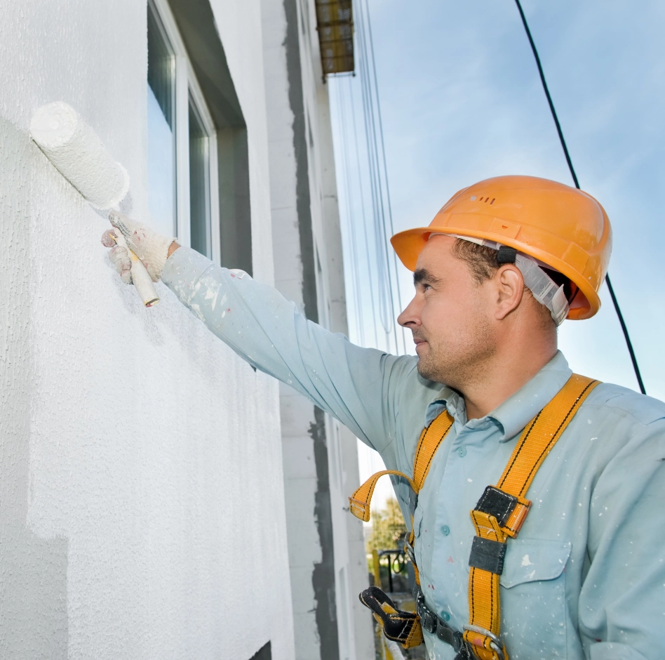A construction worker wearing an orange hard hat and safety harness is painting the exterior wall of a building white using a paint roller.