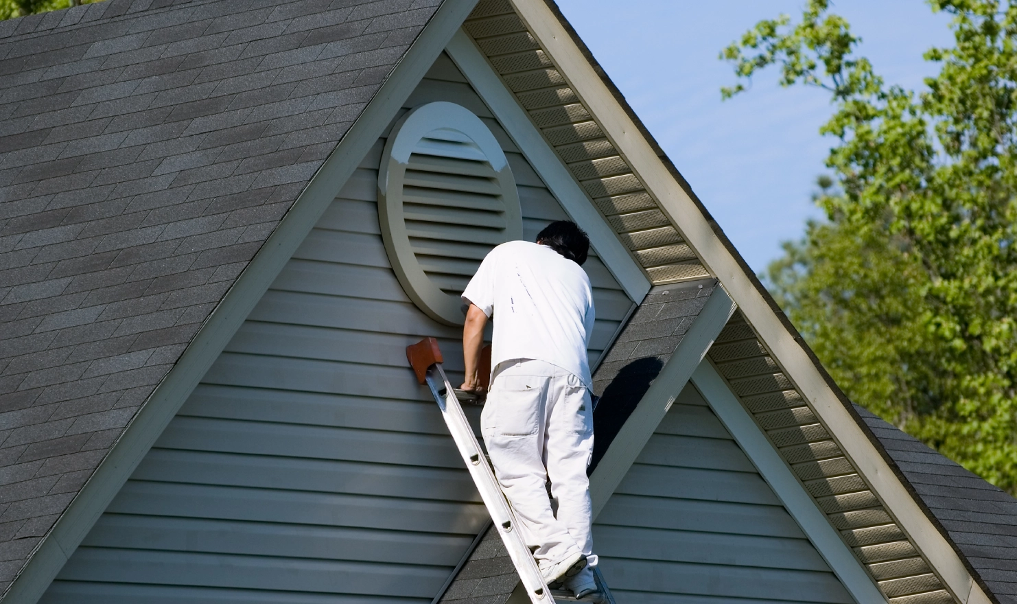 Person on ladder painting the exterior of a house gable in white, next to a round vent.