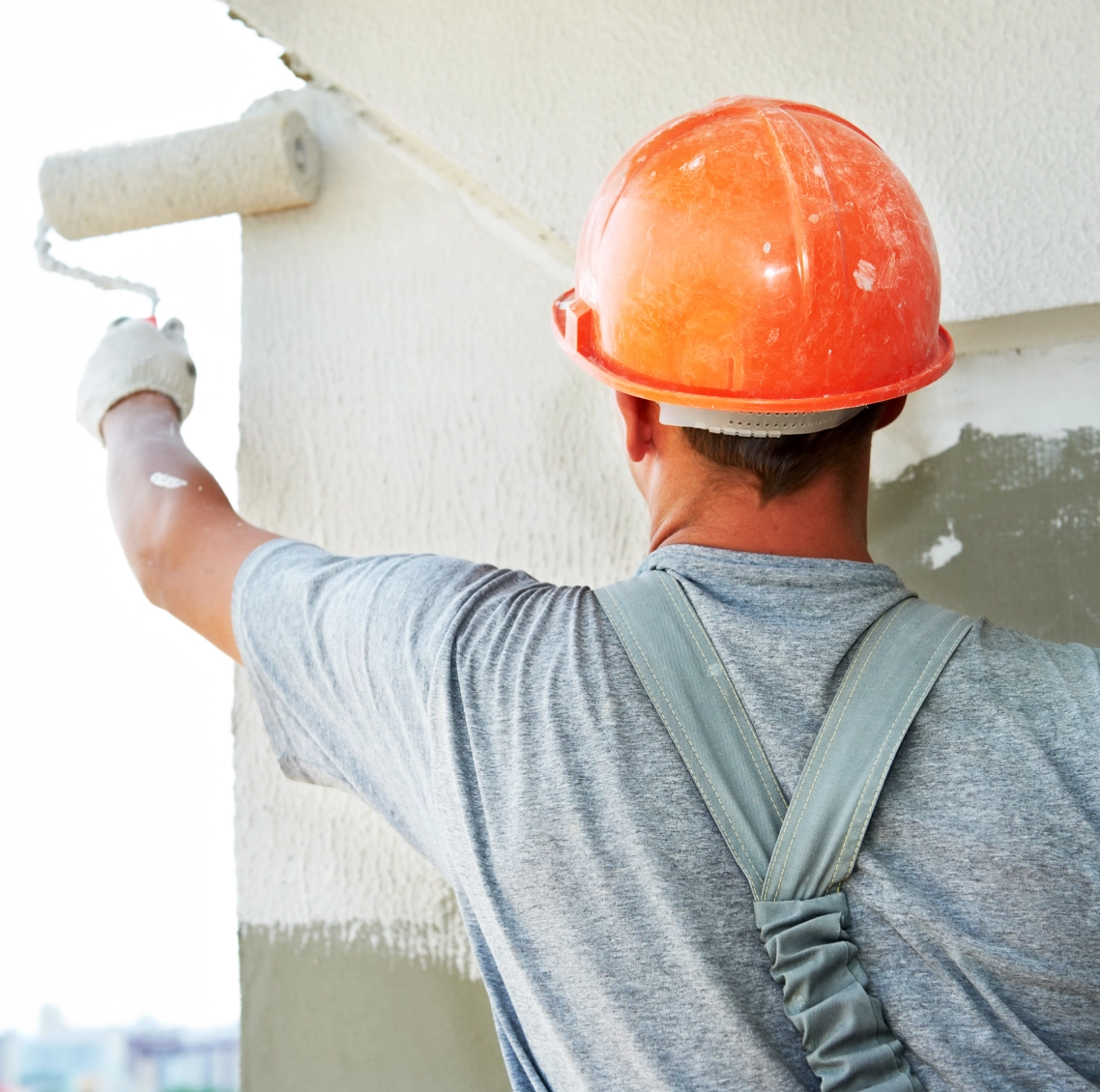 A person wearing an orange hard hat and gray shirt is painting a wall white using a roller brush.